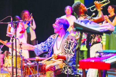 musician on drums and a trumpet player playing music, audience members standing in background clapping