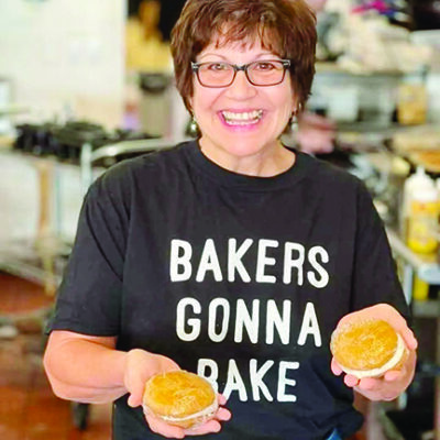 woman with short hair and glasses, wearing black shirt with words "Bakers gonna bake" holding up two wrapped vanilla whoopie pies