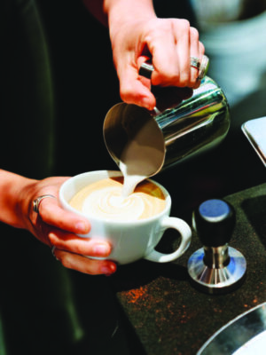 close up of hands pouring frothy milk into coffee cup to make white design on top