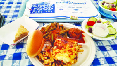 disposable plates filled with lasagna, beans, meatball, salad, and baklava sitting on blue and white checked tablecloth