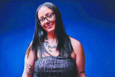 headshot of woman with long straight hair, wearing glasses, tube top, necklace with colorful stone, on blue background