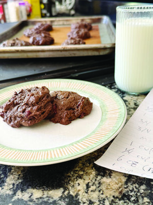 chunky chocolate cookies sitting on plate beside tall glass of milk on counter, tray of cookies in the background