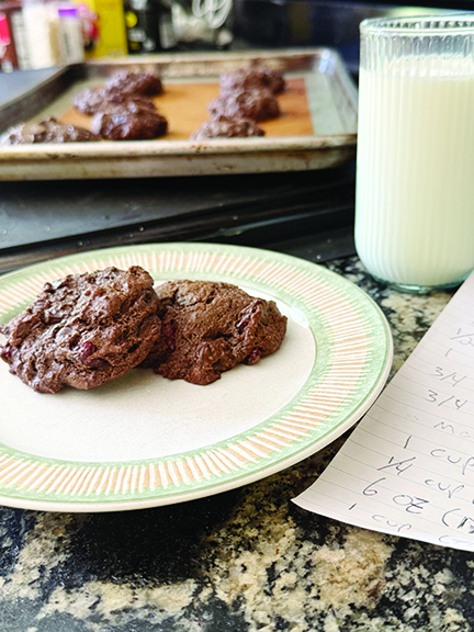 chunky chocolate cookies sitting on plate beside tall glass of milk on counter, tray of cookies in the background