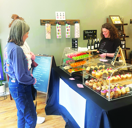 woman behind table filled with pastries and desserts for sale inside larger space, while customers look at the selection