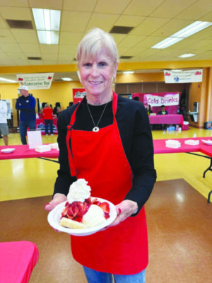 woman wearing red apron, standing in large gym space, holding dish with strawberry shortcake topped with whipped cream