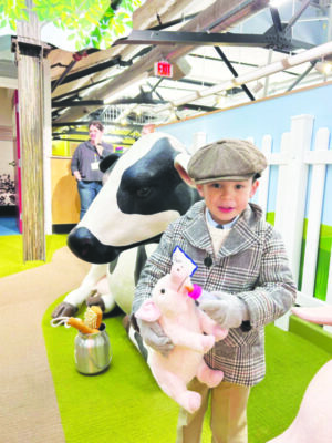 little boy wearing coat and newsboy cap, standing in colorful, child's farming exhibit, holding stuffed pig and small feeding bottle, 