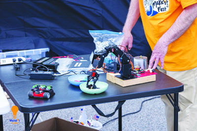 man standing at outdoor table with small robotic machine on table, long armed robot picking up something from dish
