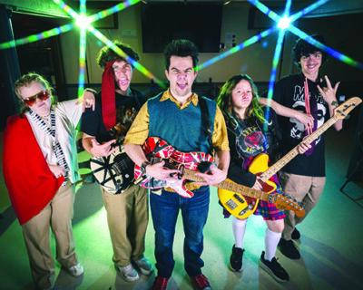 six kids holding instruments, standing on stage with rock concert lights behind them