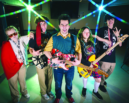 six kids holding instruments, standing on stage with rock concert lights behind them