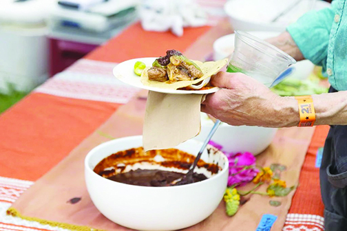 close up of elderly person's hands as they hold up plate and spoon gravy from a bowl onto meat and tortilla from outdoor table