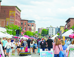 scene of street fair with people walking down the street between sidewalk tents, brick buildings rising in the background on a bright cloudy day.