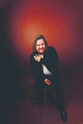 long haired man smiling, sitting on stool in dark red studio background