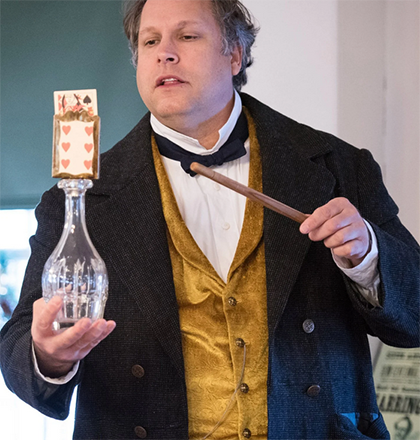 man dressed in old fashioned coat and vest, holding glass bottle with playing cards sitting on top, holding wooden wand as he does a magic trick