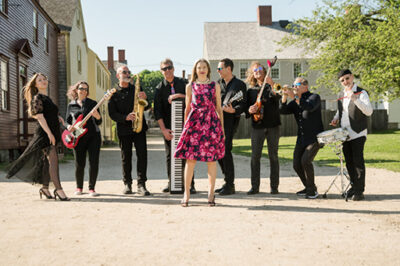 nine piece band made of women and men, standing on road outside with instruments on sunny day