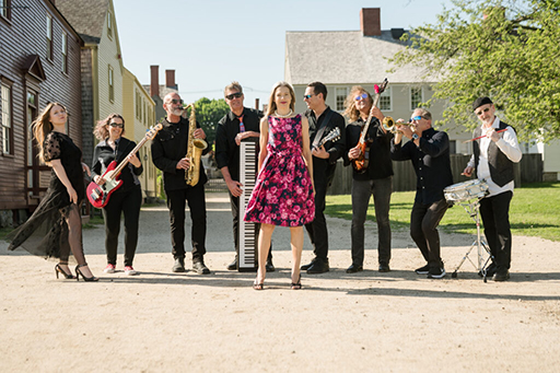 nine piece band made of women and men, standing on road outside with instruments on sunny day