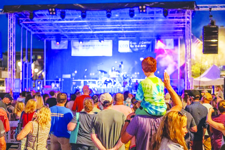 outdoor stage in evening, people standing to watch band playing, young boy sitting on man's shoulders in crowd