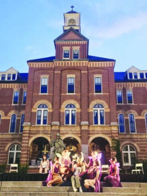 actors dressed in donkey and fairy costumes, sitting on oudoor stairs in front of brick institution building