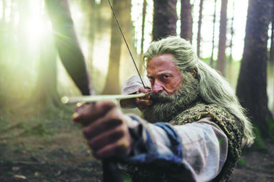 scene from The Death of Robin Hood showing Hugh Jackman with long gray hair and a beard, shooting an arrow