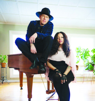 man sitting on piano in large light room, woman beside him leaning on piano