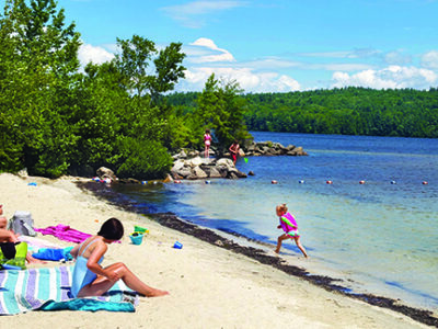 woman on blanket sitting on beach near shoreline, watching young girl running through shallow water
