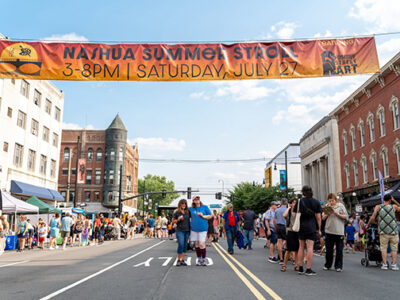 two people posing under a large banner hanging across a downtown street, crowds lining up along sidewalks at city street community event