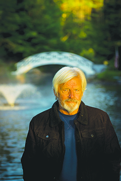 white haired and bearded man standing in front of pond and footbridge, under warm sunrise glow