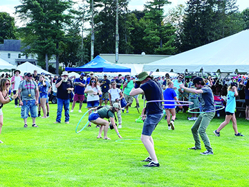 men hula hooping on grass at outdoor food festival, crowd watching, tents in background