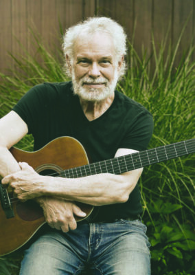 elder man with white hair and beard, sitting in front of ornamental grass and wooden fence, holding guitar