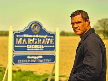 square faced man looking serious in front of town sign in Georgia field in still from Reacher