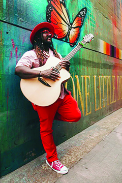 black man playing guitar, standing against wall with mural of butterfly