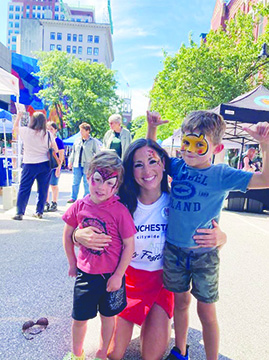 young woman crouching and side hugging two boys wearing facepaint, on downtown street on sunny day