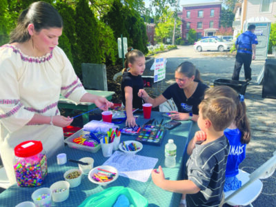 A bunch of kids surround a table with art stuff as a woman in a Greek dress helps them with their projects.