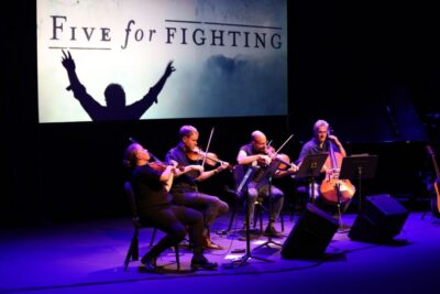 A group of violinists and chellists play on a purple-lit dark stage with the words "Five for Fighting" behind them.