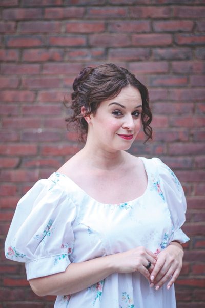A woman in a white dress gives a sly smile to the camera in front of a brick wall.