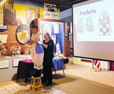 woman standing on stool wearing old fashioned bloomers and corset underwear while other women demonstrates dressing her, in museum exhibit presentation
