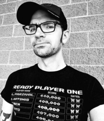 black and white image of man wearing large frame glasses and baseball cap, in front of brick wall