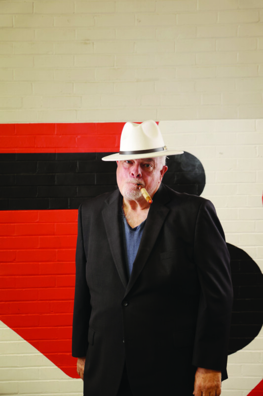 A man in a white hat and black jacket smokes a fake cigar in front of a brick wall.