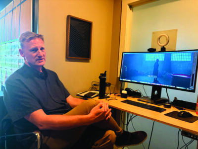 Rob Livie sits by his desk, where a monitor is running a video game.