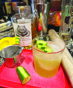 cocktail with ice and cucumbers sitting on counter with cocktail making supplies