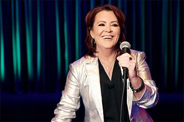 woman wearing black shirt under silver blazer holding microphone in front of green stage curtain