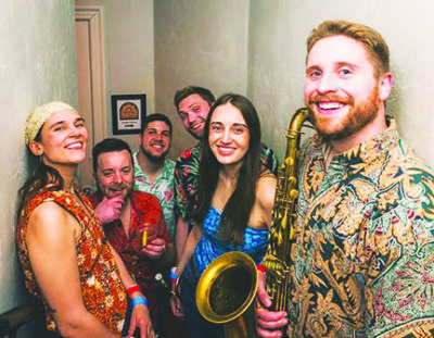 6 member band of women and men standing in hallway, one man holding saxphone