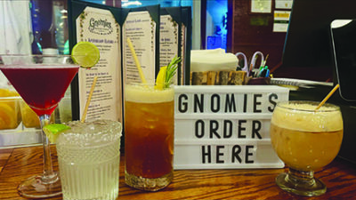 counter with sign text "Gnomies order here" surrounded by various fancy drinks in different shaped cocktail glasses